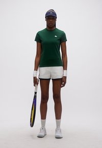 Γυναίκα που στέκεται κρατώντας ρακέτα τένις, φοράει πράσινο μπλουζάκι, λευκά σορτς, βραχιόλια καρπού, λευκά αθλητικά παπούτσια και μπλε σκούρο ζελατινοφόρο καπέλο.