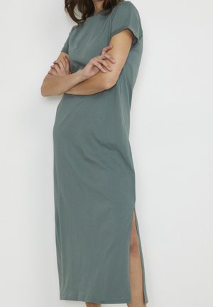 Femme portant une robe gris-vert à manches courtes, longue jusqu'aux chevilles avec une fente latérale, debout les bras croisés devant un fond blanc uni.