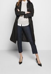 Trench-coat noir à finition brillante, superposé à un sweat-shirt gris et une chemise blanche, associé à un pantalon en denim foncé court et des escarpins noirs.