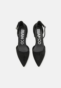 Zapatos de tacón alto de ante negro con puntera afilada, correa en el tobillo y suela plana. Logo "BIANCO" impreso en la plantilla en blanco.