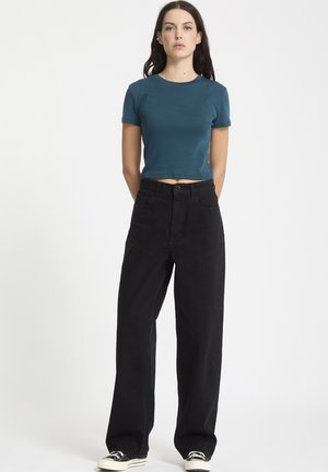 Beschnittenes türkisfarbenes T-Shirt mit kurzen Ärmeln, kombiniert mit hochtaillierten schwarzen weiten Jeans. Schwarze Sneakers runden das Outfit ab. Einfarbiger Hintergrund.