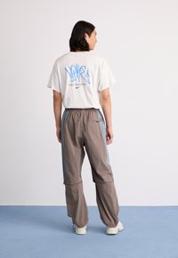 Person with shoulder-length dark hair stands facing away, wearing white graphic Nike t-shirt, gray and blue track pants, and white sneakers on blue floor.