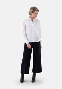 Camicia bianca a maniche lunghe con colletto, caratterizzata da bottoni sul davanti, abbinata a pantaloni neri a gamba larga e stivaletti. Tessuto liscio.