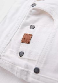 Tela de mezclilla blanca con botones oscuros y una etiqueta de cuero marrón que muestra un logotipo. La textura es suave con detalles de costura visibles.