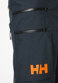 Tmavě modré látkové kalhoty mají dvě černé kapsy na zip a oranžové logo na dolní části nohavice. Textura vypadá hladce a odolně.