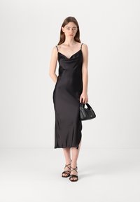 Black satin slip dress with a cowl neckline, thin straps, and a side slit. Paired with a small black handbag and black strappy sandals.