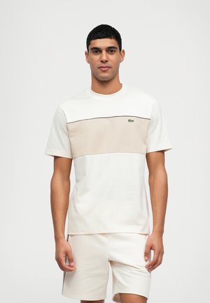 Camiseta de manga corta en algodón crema y beige con un diseño de bloques de color horizontal y un logo verde en el pecho.