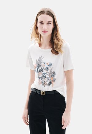 T-shirt blanc à manches courtes avec un motif floral coloré et des feuilles sur le devant, associé à un pantalon noir et une ceinture noire avec une boucle en or.