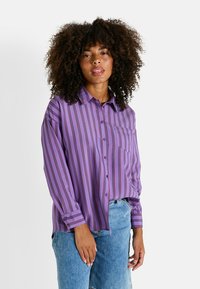 Camisa morada de botones con rayas verticales en rojo. Presenta un bolsillo delantero y mangas largas, fabricada en un tejido suave y entrelazado.