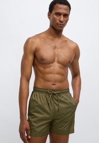 Rohelise oliivivärvi ujumispüksid, millel on nööriga vöökoht, sile tekstuur ja põlvepikkune disain ning kaks küljetaskut.