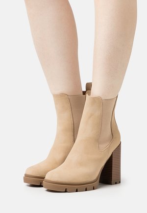 Bottines beiges en daim lisse, avec des panneaux élastiques sur les côtés et un talon épais en bois. Conçues avec un bout arrondi et une semelle crantée.