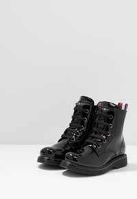 Černé kotníkové boty z lakované kůže se šněrováním, zlatými kovovými prvky a proužkovaným textilním detailem na zadní straně. Hladký, lesklý povrch.