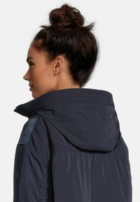 Veste matelassée bleu marine avec capuche, dotée d'un col texturé et d'accents contrastants sur les épaules. Tissu lisse et léger avec une coupe ajustée.
