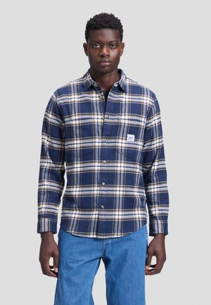 Mann trägt ein blau-weiß-beiges kariertes Hemd mit Brusttasche und hellblaue Jeans, stehend vor einem schlichten Hintergrund.
