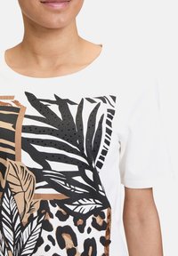 Weißes T-Shirt mit einem gemischten Druckdesign, das schwarze Blätter, Tiermuster und strukturierte Verzierungen zeigt. Kurze Ärmel und runder Halsausschnitt.