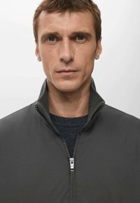 Ciemnoszara kurtka zapinana na zamek z wysokim kołnierzem, gładką fakturą i subtelnym połyskiem, noszona na ciemnym swetrze. Skupienie na dopasowanym kroju.