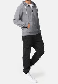 Sweat à capuche gris zippé avec cordons de serrage, poches avant, associé à un pantalon cargo noir avec poches latérales et ourlets retroussés. Porté avec des baskets blanches.