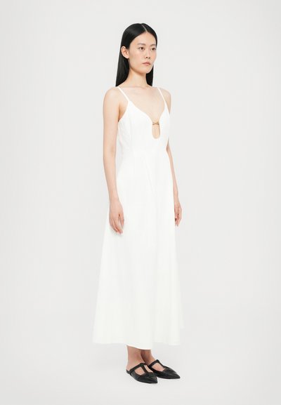 Vestido blanco sin mangas con un corte en forma de ojo de cerradura en el pecho, diseño fluido y una tela suave. Combinado con zapatos planos negros.