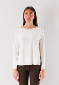 Maglione lungo crema a maniche lunghe con scollatura a barca, texture morbida e spacchi laterali, abbinato a pantaloni marroni scuri su uno sfondo neutro.