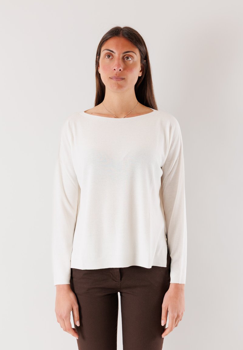 Maglione lungo crema a maniche lunghe con scollatura a barca, texture morbida e spacchi laterali, abbinato a pantaloni marroni scuri su uno sfondo neutro.