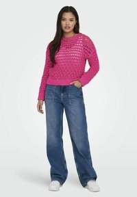 Roze gebreide trui met open weefdesign en geribbelde manchetten, gecombineerd met een wijde blauwe jeans en witte sneakers.