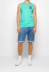 Camisa sin mangas turquesa con un bolsillo gráfico, combinada con shorts de mezclilla azul claros doblados en el dobladillo y zapatillas blancas.