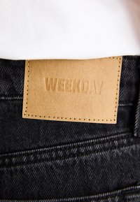 Black denim fabric with a leather patch labeled "WEEKDAY," features yellow stitching and is positioned at the waistband.