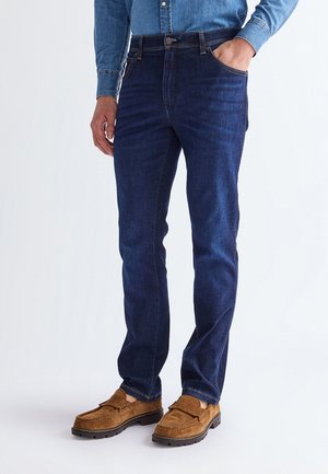 Homme portant un jean bleu foncé, une chemise en denim bleue et des mocassins en daim marron, une main dans la poche, sur fond blanc.