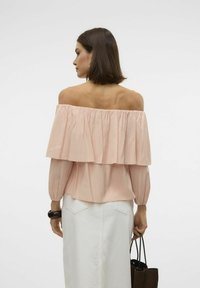 Blusa rosa off-the-shoulder con strati arricciati, maniche lunghe a sbuffo e una vestibilità rilassata. Abbinata a una gonna bianca e con una borsa nera.