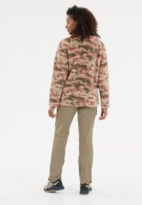 Pull en polaire beige avec un motif de camouflage rose et vert, associé à un pantalon léger beige et des chaussures de randonnée noires.