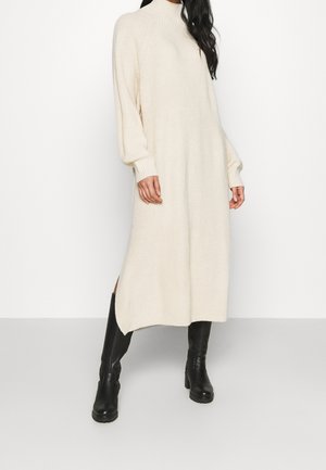 Kobieta ubrana w kremową, długą dzianinową sukienkę z długimi rękawami i czarne kozaki za kolano, stojąca na jednolitym tle.