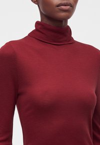 Femme portant un pull à col roulé moulant, côtelé, à manches longues de couleur rouge profond, visible du cou jusqu'au milieu du torse.
