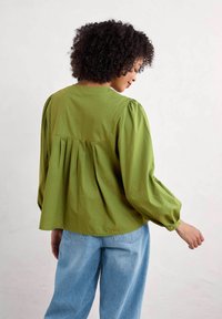 Zelená plisovaná blůza s kulatým výstřihem a nařasenými dlouhými rukávy, kombinovaná se světle modrými džínami. Hladká textura látky.