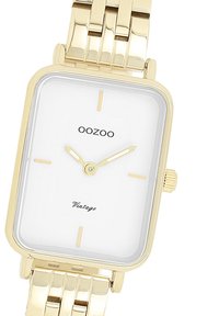 Montre-bracelet rectangulaire en or avec un cadran blanc, des index et aiguilles dorés. Elle est équipée d'un bracelet en maillons dorés et d'un logo de marque "OOZOO".