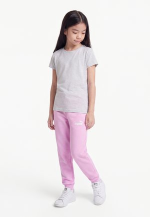 Jeune fille debout portant un t-shirt gris clair, un pantalon de jogging rose Puma et des baskets blanches sur un fond blanc uni.