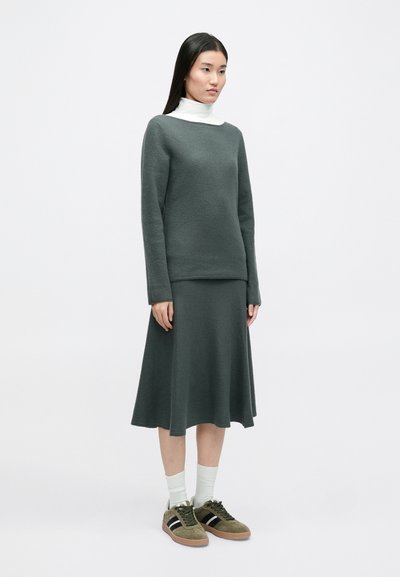 Πράσινο πουλόβερ από μείγμα μάλλινου υλικού και ταιριαστό φουστάνι με φαρδιά γραμμή, φορεμένο με λευκό ζιβάγκο και πράσινα αθλητικά παπούτσια με μαύρες ρίγες. Λείος υφής.