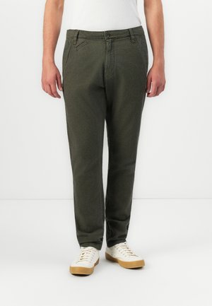 Man wearing dark green casual trousers and white sneakers standing against a plain white background.