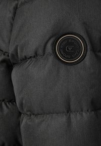 Sort dunjakkeafsnit med tekstureret stof, synligt syning og en cirkulær sort logopatch med beige kant.