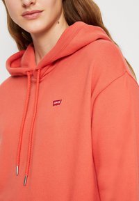 Coral pink hoodie with a drawstring and silver tips, featuring a small red Levi's patch on the chest. Soft fabric and relaxed fit.