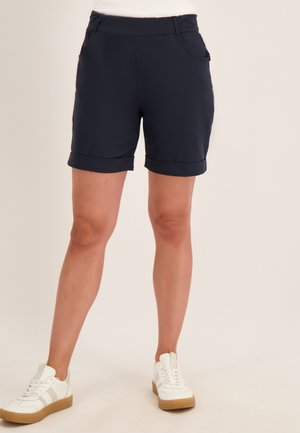 Persoon die zwarte omslagshorts en witte sneakers met beige zolen draagt, staand tegen een egale lichte achtergrond.