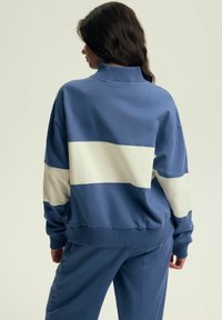 Niebieska bluza z wysokim kołnierzem, z szerokim, off-white poziomym pasem na plecach. Miękka tkanina i ściągacze przy mankietach.