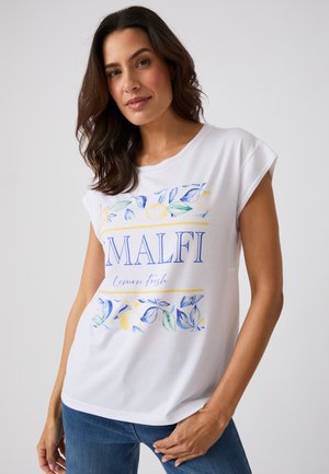 Frau mit dunklem Haar, die ein weißes ärmelloses Oberteil mit blauem und gelbem Zitronenprint und dem Text "MALFI Lemon fresh," trägt, kombiniert mit blauen Jeans.