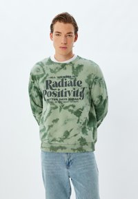 Grön tie-dye sweatshirt med ribbade muddar och nederkant, med fet svart text: "Stråla Positivitet" och ytterligare motiverande fraser.