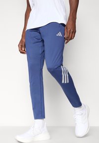 Μπλε αθλητικό παντελόνι κατασκευασμένο από λείο, ελαστικό ύφασμα, με γκρι λεπτομέρειες τριών ρίγων στα πλάγια και σχεδιασμό με στενό τελείωμα στα πόδια.