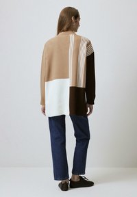 Gehaakte trui met bruine, beige en witte secties, voorzien van verticale strepen, een relaxed fit, gecombineerd met donkere spijkerbroek en bruine schoenen.