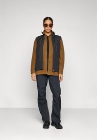 Zwarte mouwloze vest over een bruine overhemd met knopen, gecombineerd met donkere broek en zwarte schoenen. Zonnebril maakt de outfit compleet.