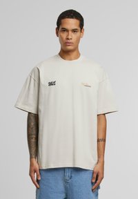Beige oversized t-skjorte med svart tekst "out of office" på venstre bryst og oransje tekst på høyre side. Korte ermer og rund hals.