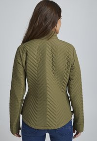 Veste matelassée vert olive avec un motif en chevrons, col montant et manches longues, associée à un jean en denim.