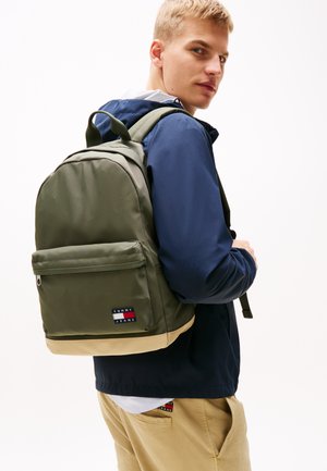Mochila verde oliva con respaldo acolchado, base beige y bolsillo frontal con cremallera que presenta una etiqueta negra. Correas ajustables y un asa superior.