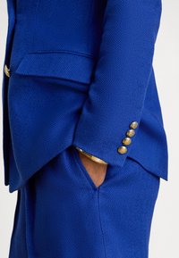 Modra blazer iz tkanine z ribjo kostjo z zlato gumbi, z izdelano krojenjem in stranskim žepom. Tekstura je gladka, s podrobnim šivanjem.